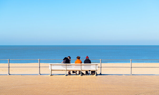Waterfront promenade bench with three people sitting in front of Oostende (Ostend) North Sea beach, West Flanders, Belgium.