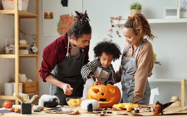Happy african american family mother, father and child son carving pumpkin for Halloween holiday together at home