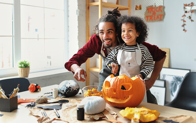 Ethnic father removing pulp from ripe pumpkin while carving jack o lantern with little son for...