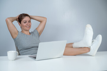 Caucasian woman lifted her leg with plaster to work desk and works on laptop on white background