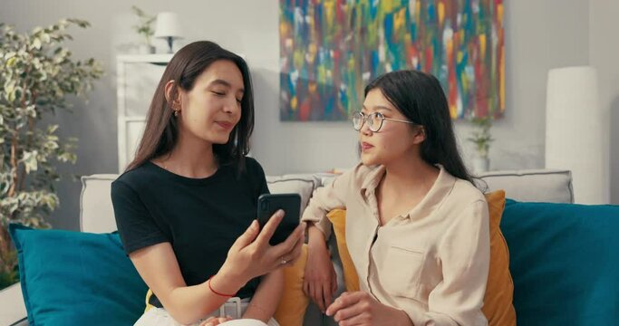 Two pretty best friends of Asian Korean beauty spend an afternoon on the living room couch, talking, the woman shows vacation photos on her phone, they look together, smile, comment