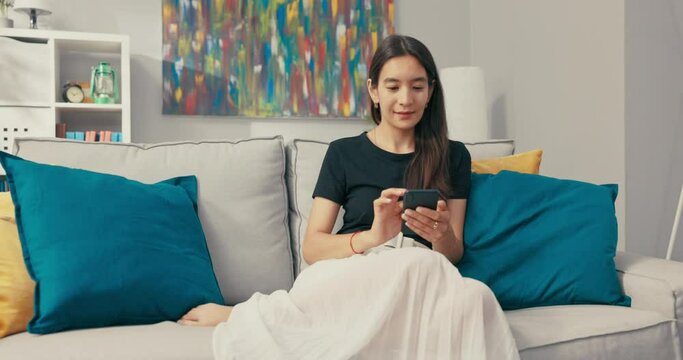 A beautiful Asian woman with brunette hair sits smiling gently on a couch in living room, dressed in linen skirt and loose black tshirt, holding phone in hands, texting with friends, chatting