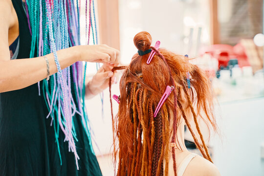 Red-haired girl makes fashionable braids dreadlocks. Safe removable dreadlocks from kanekalon. Cool hairstyle made of artificial hair. Hairstyle of rainbow Afro-pigtails.