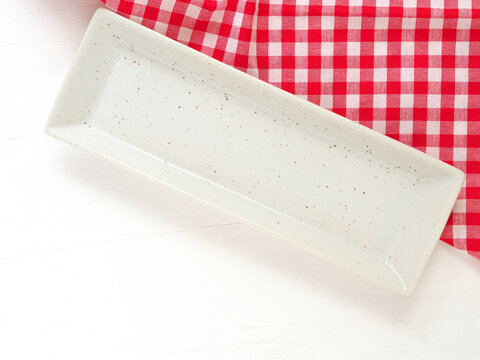 Top view, Empty long rectangle white plate with brown dot on red and white checkered fabric napkin on white table background. Concept kitchen utensils and tableware. flat lay with copy space..