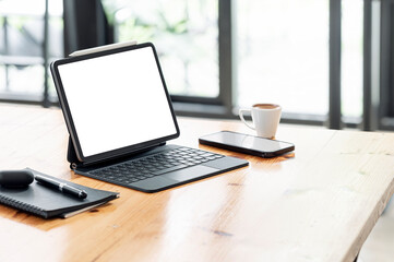 Mockup blank white screen tablet with magic keyboard and gadget on wooden table in cafe room.
