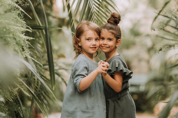 Two cute smiling little girls belonging to different races, in linen clothes, holding hands and walking in the botanical garden. Children explore tropical plants and flowers in the greenhouse.