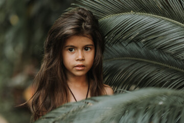The face of a little girl surrounded by tropical leaves. Closeup portrait of a beautiful swarthy...
