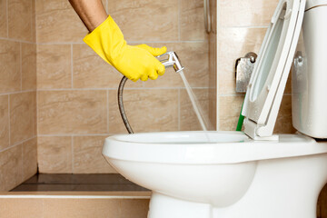 Human hand in yellow rubber glove are using a hose to spray water to clean the toilet bowl.