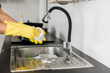 Close up human hands in yellow rubber gloves washing a ceramic coffee mug in the kitchen sink.