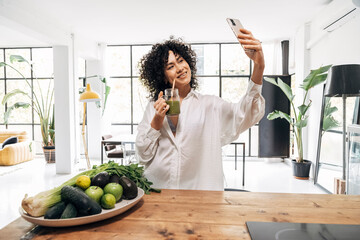 Young smiling african american woman takes selfie drinking green juice reusable bamboo straw in bright loft apartment