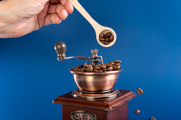 A hand sprinkles coffee beans into a manual coffee grinder with a spoon. Dark blue background. Photo.