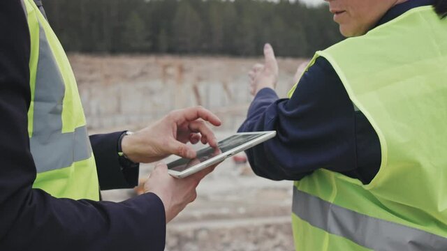 Mid-section slowmo of unrecognizable quarry manager with tablet discussing work with female supervisor while standing by granite quarry