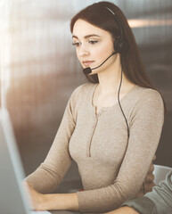 Young friendly girl in headsets is talking to a firm's client, while sitting at the desk in sunny office. Call center operators at work