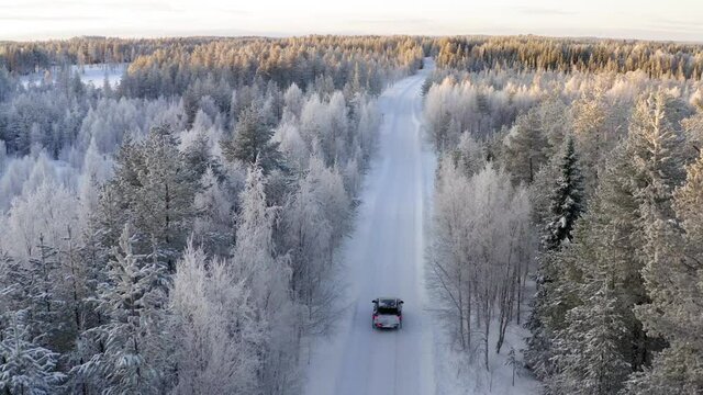 Aerial view of car driving on white wild forest path passing tall trees covered with ice and snow, winter equipment on jeep outlander for crossing rural environment on north
