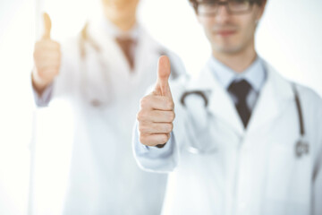 Two male doctors standing as a team and showing thumbs up as a symbol of the best service for patients in sunny clinic. Medicine concept