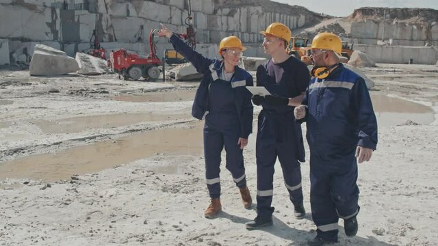 Slowmo tracking of female and male workers in uniform and hard hats walking in granite quarry and talking while looking at tablet