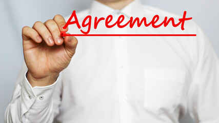 A businessman in a white shirt writes a text AGREEMENT on a white background with a red marker . business concept