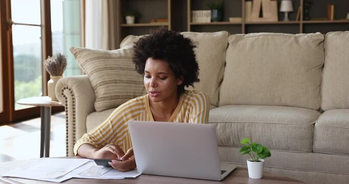African woman sit in living room makes calculations on calculator, pay household bills and loan through e-bank application on laptop. Personal family budget management, modern tech, accounting concept