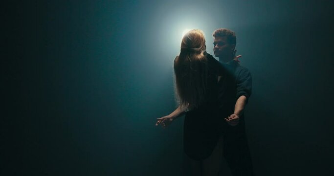 Attractive blonde seduces handsome partner in dance, flirtation in movements, woman leans back swinging her hair, delicacy, femininity, romantic atmosphere, dark scene lit by strong lamp