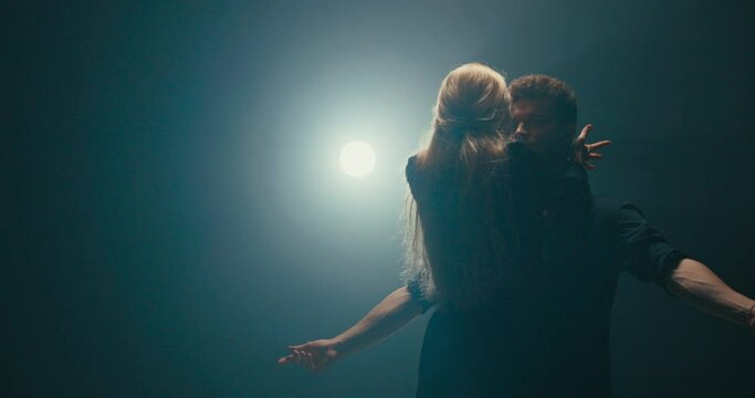 Attractive blonde seduces handsome partner in dance, flirtation in movements, woman leans back swinging her hair, delicacy, femininity, romantic atmosphere, dark scene lit by strong lamp