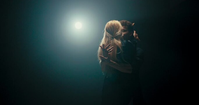 Attractive blonde seduces handsome partner in dance, flirtation in movements, woman leans back swinging her hair, delicacy, femininity, romantic atmosphere, dark scene lit by strong lamp