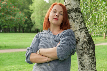 Attractive young woman with red hair in a blue knitted sweater stands by a birch tree and looking at the camera haughtily. Overconfident woman folded her arms across her chesty