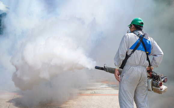 Rear view of healthcare worker in protective clothing using fogging machine spraying chemical to eliminate mosquitoes at general location in community