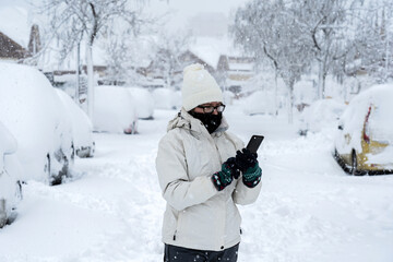 Fototapeta na wymiar mujer con gorro de lana y abrigo blanco mirando el móvil con una tremenda nevada en la calle