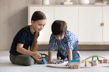 Happy friendly adorable little brother sister kids sitting on cozy floor carpet, playing with toy railway road, enjoying stress free leisure weekend playtime together at home, daycare concept.