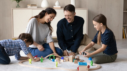 Fototapeta premium Happy young hispanic couple parents having fun with little adorable kids son daughter playing wooden toys railway road, sitting together on floor carpet, joyful family entertaining in living room.