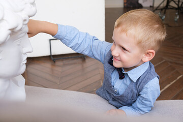 Portrait of a five-year-old boy in profile. Boy touches a plaster bust while lying on the couch. Boy at creative lesson in an art workshop. Child studies the structure of face using plaster figure.