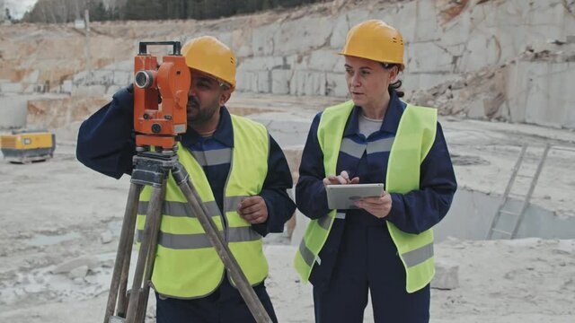 Slowmo tracking of Arab male granite quarry worker in vest and hard hat using surveying measuring equipment and talking to female colleague making notes on tablet