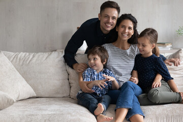 Fototapeta premium Portrait of cheerful friendly latin hispanic caucasian family with small adorable kids son daughter sitting on cozy couch, copy space for advertisement text, real estate, home ownership concept.