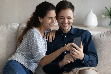 Laughing beautiful millennial latin married couple using cellphone, choosing goods in internet...