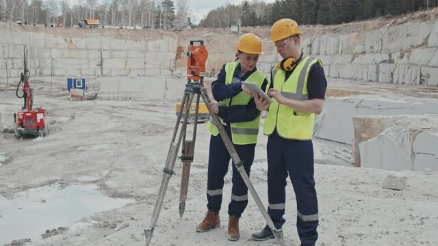 Slowmo tracking of professionals working in granite quarry. Female worker in vest and hard hat using surveying measuring equipment and talking to male colleague making notes on tablet