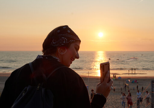 A young smiling female taking a photo of a sunset on a crowded seashore