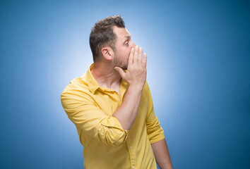 Young man holding hand near mouth and telling secret over blue background, dresses in yellow shirt. Gossip concept