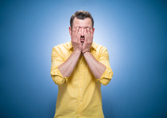 Tired guy over blue background, dresses in yellow shirt. Sleepy man rubbing her eyes. Young man covers face. Sleepy expression
