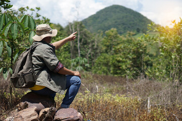 Naklejka na ściany i meble Side view of male hiker sits on stone and point something at the mountain and forest. Concept : Eco traveling. Recreation on holiday. Trekking in the forest alone. Survival in forest. Copy space 
