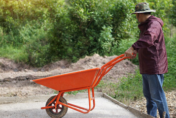 Asian man push orange wheel barrow cart trolley that use for construction to carry cement mixture at worksite. Concept : Construction worker's  tool.     