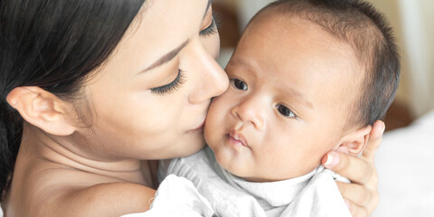 Portrait of enjoy happy love family asian mother playing with adorable little asian baby.Mom kiss with cute son moments good time in a white bedroom.Love of family concept