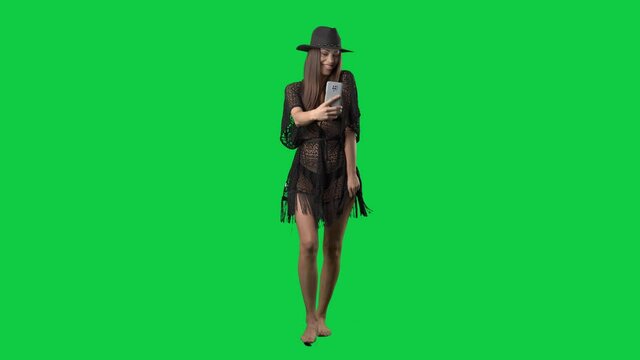 Walking barefoot young bikini woman with hat taking selfies with smartphone. Full body on green screen chroma key background