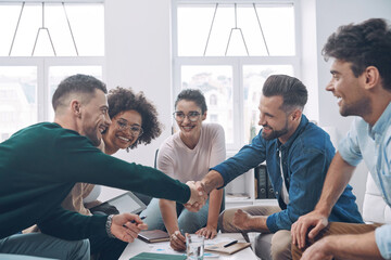 Two handsome young men shaking hands and smiling while having meeting with colleagues in office