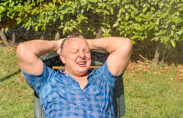 Portrait of happy mature man under splashing water on a hot day in the garden.