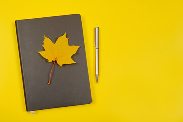 On the gray notebook is an autumn yellow maple leaf and a pen on a yellow background.The concept of teacher's day, things for businessmen, education, top view, autumn planning.Copyspace