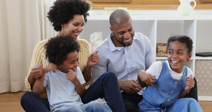 Beautiful cheerful mixed race African couple tickling little son and pre-teen daughter sit together on warm floor in modern play room. Happy homeowners family, weekend leisure, bond and ties concept