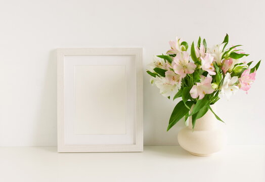 White wooden frame mockup on white table, ceramic vase with pink and white flowers bouquet near white wall background. Minimal interior. Copy space.