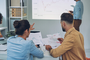 Obraz premium Group of young modern people in smart casual wear analyzing data on projection screen in the office