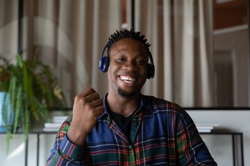 Head shot of millennial Black hipster guy in headphones making video call, looking, talking at...