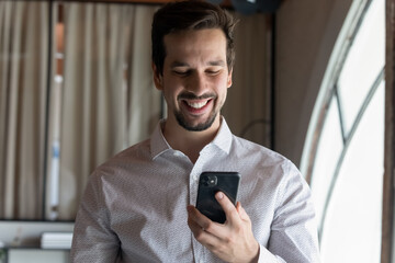 Happy confident millennial businessman reading text message on online chat, making virtual call, getting good news, looking at mobile phone screen, smiling, laughing, using banking app on smartphone
